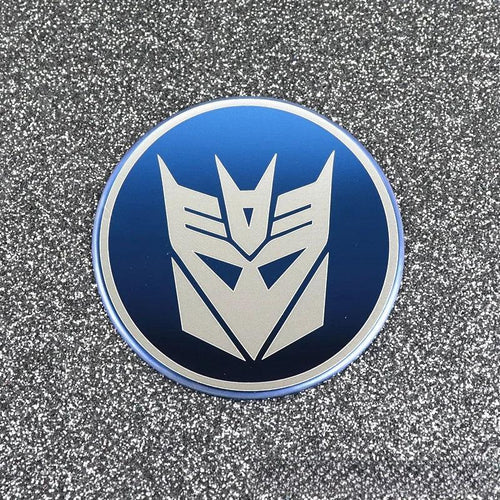 Badge rond logo Decepticon argenté sur fond bleu métallique, décor pailleté – Transformers.