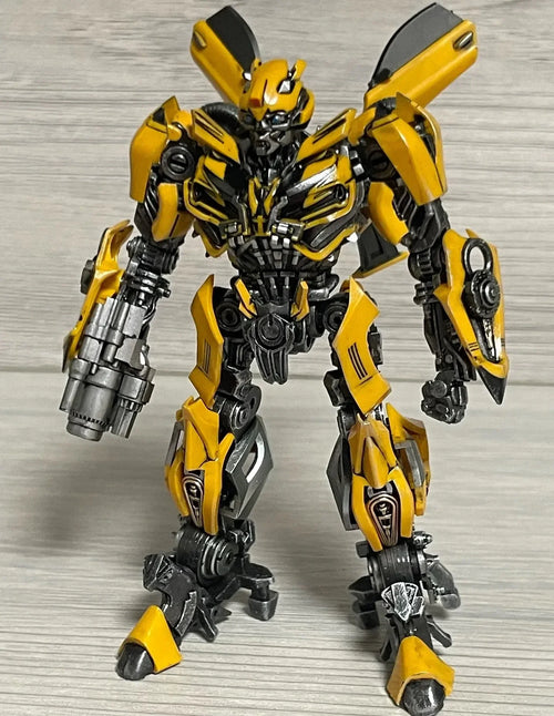 Figurine robot articulée jaune et grise debout, fond bois clair, style mécanique détaillé – Transformers.